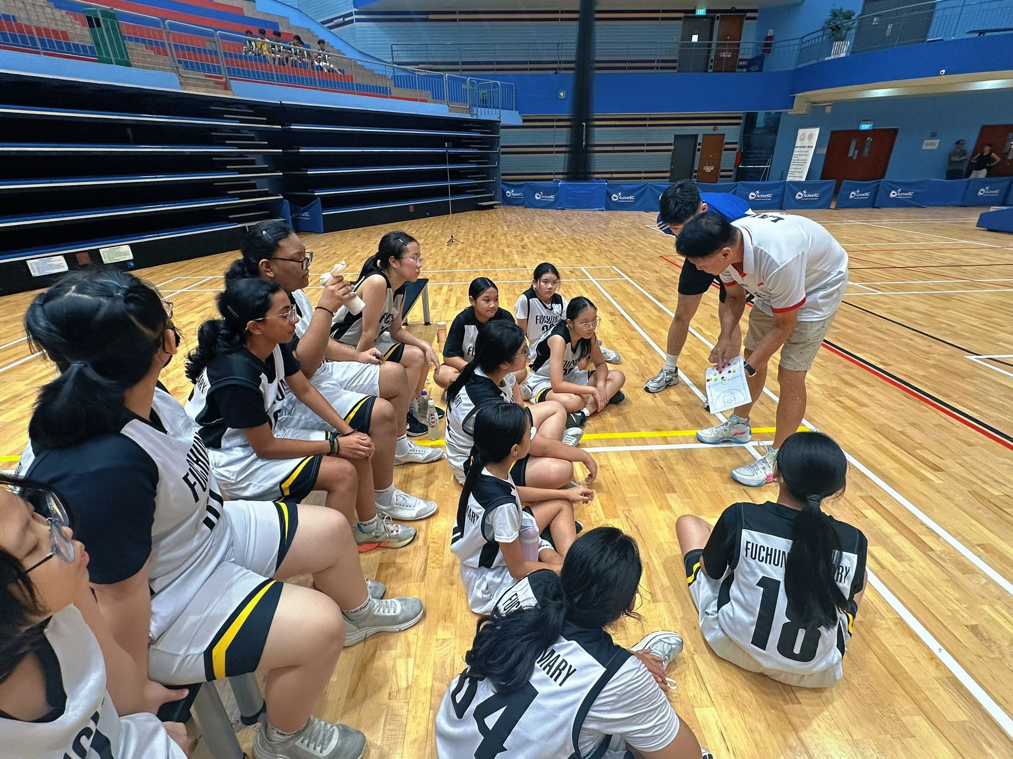 Basketball coach briefing the Fuchun Primary School girls' basketball team during a match at an indoor sports hall.
